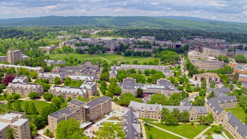 aerial view of Virginia Tech drillfield and surrounding buildings on a sunny day. Green mountains and horizon are in the background.